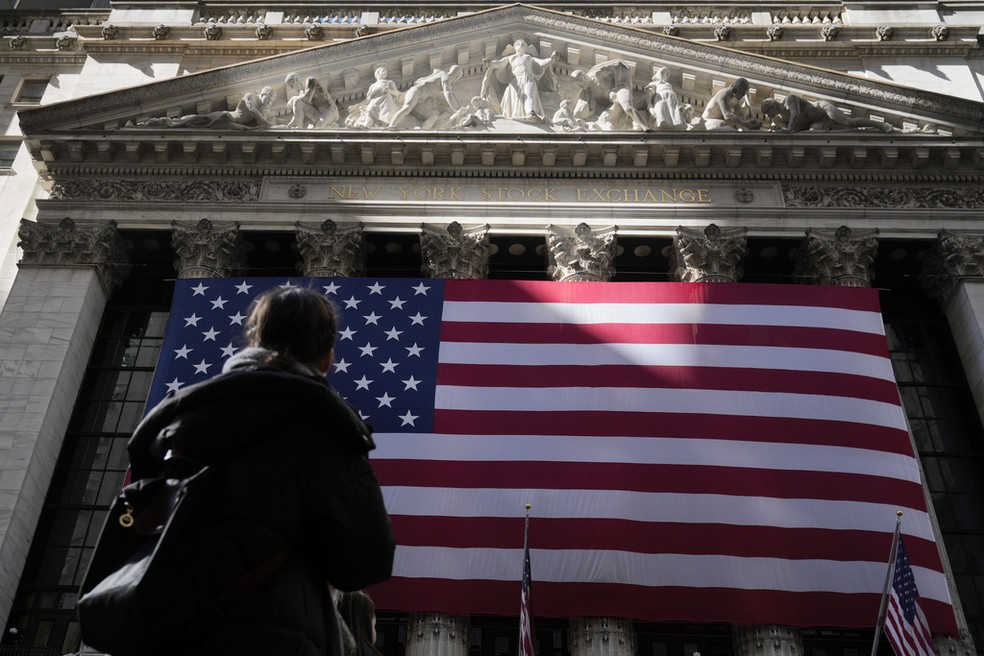 Mulher olha para bandeira dos Estados Unidos pendura em prédio da Bolsa de Valores de Nova York， em 26 de fevereiro de 2025. — Foto: Seth Wenig/AP